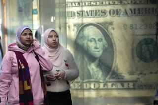 FILE - Egyptians walk past a poster depicting a U.S. dollar outside an exchange office in Cairo, Egypt, Thursday, Feb. 9, 2023. Egypt’s annual inflation rate set a record high in June, as the most populous Arab country continues to battle price hikes and a depreciating currency, the Egyptian statistics bureau said Monday, July 10, 2023. (AP Photo/Amr Nabil, File)