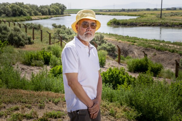Ken Mochizuki, 69, whose parents were incarcerated at Minidoka, poses in front of reconstructed barbed wire fencing near the entrance at Minidoka National Historic Site, Saturday, July 9, 2023, in Jerome, Idaho. Mochizuki, who first began attending pilgrimages as a journalist for the International Examiner in Seattle, has authored several books on the camps. His parents never returned to the site or discussed their time at Minidoka, leading Mochizuki to a "lifetime of reconstructing" what the experience was like for them. (AP Photo/Lindsey Wasson)