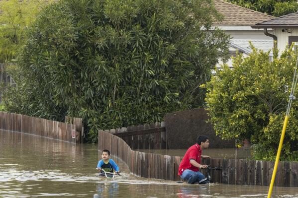 A boy and a man ride bicycles through floodwaters in Watsonville, Calif., Saturday, March 11, 2023. (AP Photo/Nic Coury)