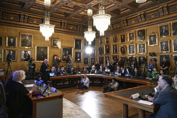 Mats Larsson, member of the Royal Academy of Sciences, standing at left, speaks during the announcement of the winner of the 2023 Nobel Prize in Physics, at the Royal Academy of Sciences, in Stockholm, Tuesday, Oct. 3, 2023. The Nobel Prize in physics has been awarded to Pierre Agostini, Ferenc Krausz and Anne L’Huillier for looking at electrons in atoms by the tiniest of split seconds. (Anders Wiklund/TT News Agency via AP)
