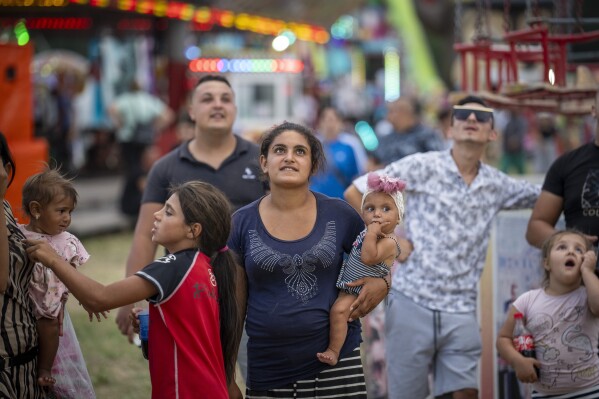 A woman holds a baby while looking along with others at people enjoying rides at a fair in Hagioaica, Romania, Thursday, Sept. 14, 2023. For many families in poorer areas of the country, Romania's autumn fairs, like the Titu Fair, are one of the very few still affordable entertainment events of the year. (AP Photo/Vadim Ghirda)