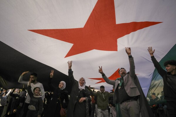Syrians walk beneath a "revolutionary" Syrian flag during a celebratory demonstration following the first Friday prayers since Bashar Assad's ouster, in Damascus' central square, Syria, on Friday, Dec. 13, 2024. (AP Photo/Leo Correa)