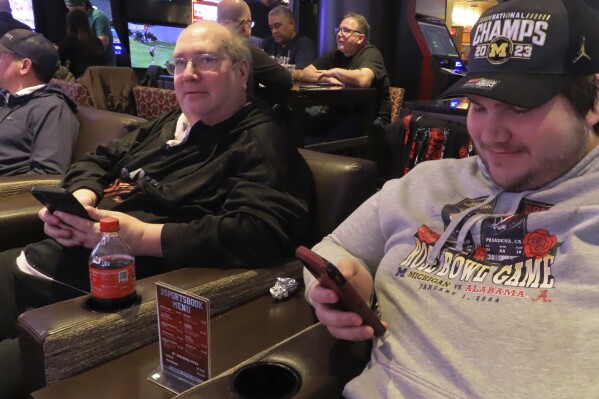 Mark F. Bawers, left, and his won Mark A. Bawers, right, check their phones while waiting for the NCAA basketball tournament to begin as they sit in the sports book at Golden Nugget casino in Atlantic City N.J. on Wednesday, March 21, 2024. The American Gaming Association estimates Americans will wager $2.72 billion with legal outlets this year. (AP Photo/Wayne Parry)
