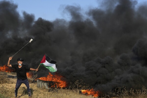 A Palestinian protester uses sling shots to hurls stones while waves Palestinian flag as others burn tires during clashes with Israeli security forces along the frontier with Israel, east of Gaza City, Friday, Sept. 22, 2023. The Israeli military said it struck three posts belonging to Hamas, the Islamic militant group that has controlled Gaza since 2007, following a number of incendiary balloons launched from Gaza into Israel. This is the latest violence to roil the territory as Palestinians stage routine protests by the border fence. (AP Photo/Adel Hana)