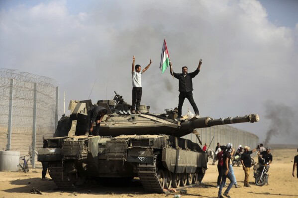 Palestinians wave their national flag and celebrate by a destroyed Israeli tank at the southern Gaza Strip fence east of Khan Younis on Saturday, Oct. 7, 2023. (APPhoto/Yousef Masoud)