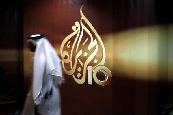 FILE - A Qatari employee of Al-Jazeera Arabic language TV news channel walks past the logo of Al-Jazeera in Doha, Qatar. The pan-Arab news network Al Jazeera has condemned a recent decision by Egyptian authorities to brand some of its journalists as terrorists. The media outlet, which is owned by the Gulf state of Qatar, said that “a number” of its Egyptian journalists and presenters had been added to a list of alleged terrorists published in an official newspaper earlier this month following a ruling by the Cairo Criminal Court. (AP Photo/Kamran Jebreili, File)