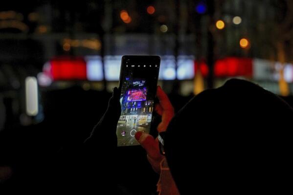 A man uses his phone to film a screen outside a mall showing a live broadcast of the fireworks explode over the iconic Bird's Nest Stadium during the closing ceremony of the 2022 Winter Olympics, in Beijing, Sunday, Feb. 20, 2022. (AP Photo/Andy Wong)