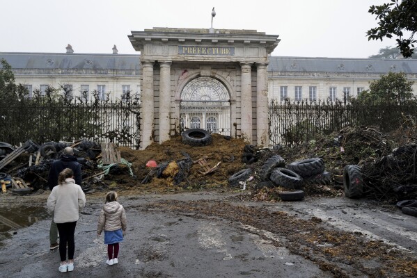 FILE - People look on at slurry, fertilizer and tires dumped by farmers at the entrance to the provincial government building in Agen, southwest France, on Saturday, Jan. 27, 2024. French farmers have vowed to continue their protests and are maintaining traffic barricades on some roads. of the country's main roads. The government announced a series of measures on Friday, January 26, 2024, but farmers claim these do not fully meet their demands.  (AP Photo/Fred Scheiber, File)
