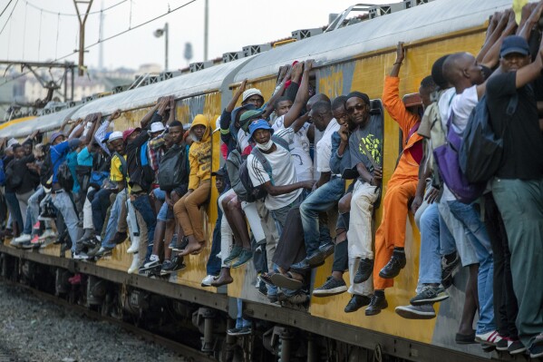 FILE - Train commuters hold on to the side of an overcrowded passenger train in Soweto, South Africa. Monday, March 16, 2020. (AP Photo/Themba Hadebe, File)