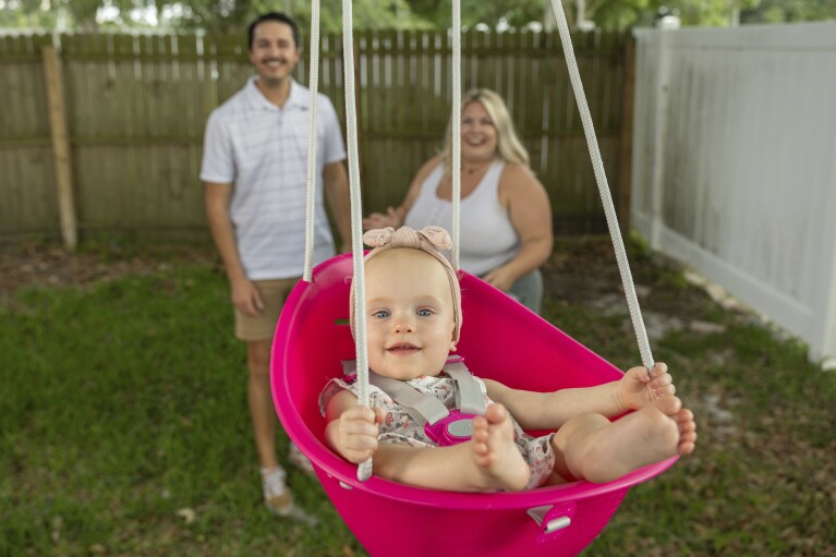 Sam Earle, left, and his wife, Tori, watch their daughter, Novalie, swing in their backyard Tuesday, May 7, 2024, in Lakeland, Fla. Novalie was born through an embryo adoption. “God can use everything to His glory,” says Sam, 30. “There’s certainly an aspect that you consider with IVF: the ethics of freezing more embryos than you need. … But for families who struggle with infertility, it’s a beautiful opportunity.” (AP Photo/Mike Carlson)
