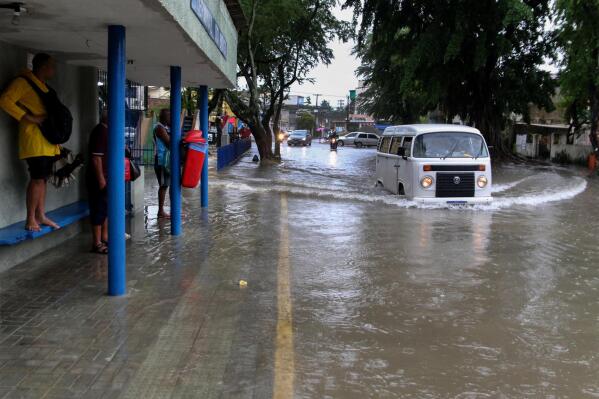 A woman stands on a bus stop bench as a driver of a Volkswagen van navigates a flooded street in Recife, state of Pernambuco, Brazil, Saturday, May 28, 2022. (AP Photo/Marlon Costa/Futura Press)