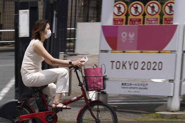 A woman wearing a protective mask to help curb the spread of the coronavirus rides a bicycle past a banner for the Tokyo 2020 Olympic and Paralympic Games Wednesday, May 26, 2021, in Tokyo. (AP Photo/Eugene Hoshiko)