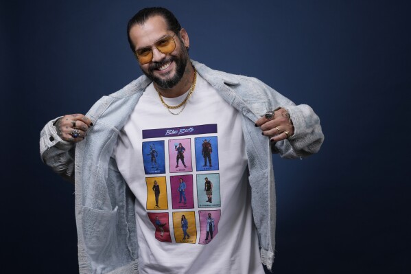 Angel Manuel Soto, director of "Blue Beetle." shows off a T-shirt displaying the film's cast as he poses for a portrait, Friday, Aug. 4, 2023, at the London Hotel in West Hollywood, Calif. The "Blue Beetle" cast members could not promote the film as per SAG-AFTRA guidelines during the actors' strike. (AP Photo/Chris Pizzello)