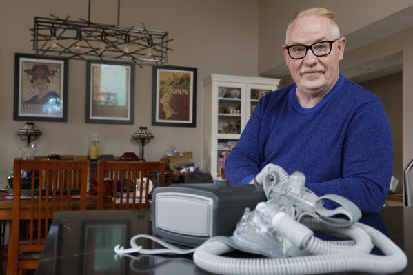 FILE - Jeffrey Reed, who experienced persistent sinus infections and two bouts of pneumonia while using a Philips CPAP machine, poses with the device at his home, Oct. 20, 2022, in Marysville, Ohio. The company responsible for a global recall of sleep apnea machines will be barred from resuming production at U.S. facilities until it meets a number of safety requirements under a long-awaited settlement announced Tuesday, April 9, 2024 by federal officials. (AP Photo/Jay LaPrete, file)