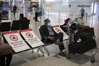 A man sits wearing a face shield in the arrivals area on the first day of new rules that people arriving in Britain from overseas will have to quarantine themselves for 14 days to help stop the spread of coronavirus, at Heathrow Airpot in London, Monday, June 8, 2020. The British government has said that anyone caught not complying with the quarantine will face a fine. (AP Photo/Matt Dunham)