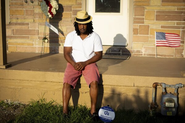 Dion Green sits on his front porch on Tuesday, Aug. 6, 2019 in Dayton, Ohio, with a flower arrangement and an American flag honoring his father, Derrick Fudge, who was killed in Sunday’s mass shooting in Dayton.   Green just wanted to have some fun with his family in downtown Dayton after what had been a tough couple of months in the aftermath of damaging tornadoes. But his Saturday night out ended tragically, with his father dying in his arms, his eyes looking into his as he took his final breath.   (AP Photo/Robert Bumsted)