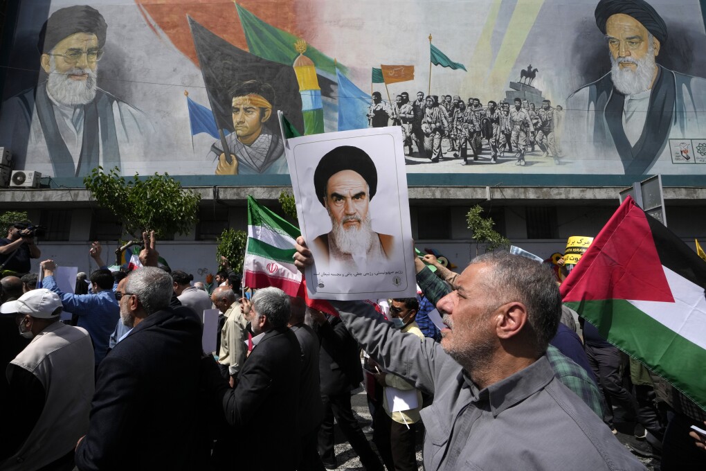 FILE - Iranian worshippers walk past a mural showing the late revolutionary founder Ayatollah Khomeini, right, Supreme Leader Ayatollah Ali Khamenei, left, and Basij paramilitary force, as they hold posters of Ayatollah Khomeini and Iranian and Palestinian flags in an anti-Israeli gathering after Friday prayers in Tehran, Iran, April 19, 2024. (AP Photo/Vahid Salemi, File)