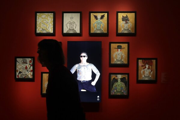 In this Wednesday, July 10, 2019 photo, a woman walks past an interactive display of artwork and tattoos during a media preview of "Ed Hardy: Deeper than Skin" at the de Young Museum in San Francisco.  The new exhibition sheds light on Hardy as a prolific artist and tattoo pioneer. It contains 300 paintings, drawings, prints and objects that track Hardy's  evolution. (AP Photo/Jeff Chiu)