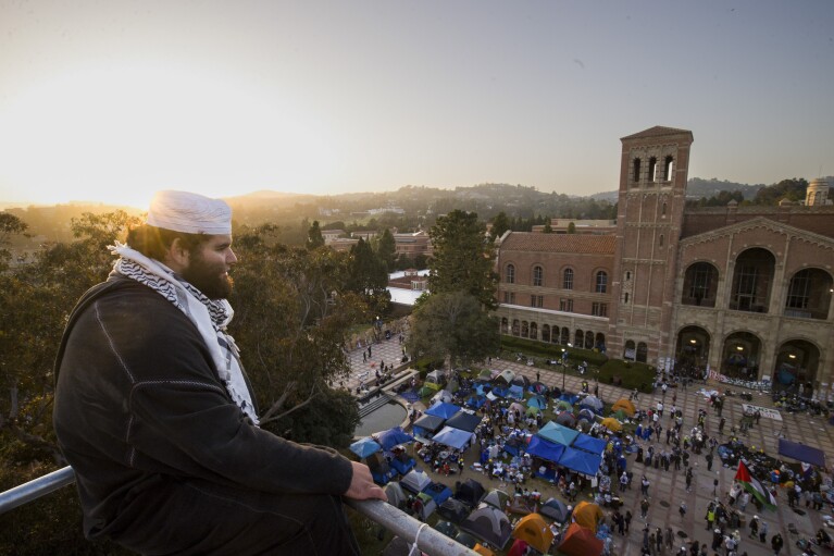 Um manifestante olha para um acampamento a partir de andaimes de construção no campus da UCLA após confrontos entre grupos pró-Israel e pró-Palestina, quarta-feira, 1º de maio de 2024, em Los Angeles.  (Foto AP/Ethan Swope)
