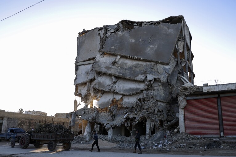 People walk next to the damage of a building that was destroyed in the February 2023 earthquake, in Idlib province, Syria, Saturday, Jan. 27, 2024. A year after the devastating 7.8 magnitude earthquake struck southern Turkey and northwestern Syria, a massive rebuilding effort is still trudging along. The quake caused widespread destruction and the loss of over 59,000 lives. (AP Photo/Omar Albam)