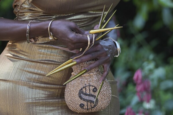A racegoer in a gold dress with long gold nails and a dollar symbol handbag poses for a photograph on the third day of the Royal Ascot horse race meeting, traditionally called Ladies Day, at Ascot, England, Thursday, June 19, 2025. (AP Photo/Kin Cheung)
