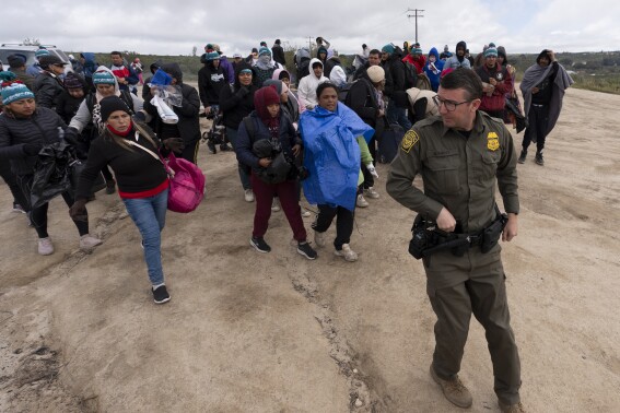 FILE - People seeking asylum, including a group from Peru, walk behind a Border Patrol agent towards a van to be processed after crossing the border with Mexico nearby, on April 25, 2024, in Boulevard, Calif. President Joe Biden has ordered a halt to asylum processing at the U.S. border with Mexico when arrests for illegal entry top 2,500 a day, which was triggered immediately. (AP Photo/Gregory Bull, File)