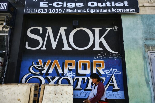 In this Wednesday, Aug. 28, 2019, photo, a woman smokes on the street next to a wholesale vape shop in downtown Los Angeles. A short walk from police headquarters in the heart of downtown, a cluster of bustling shops are openly selling packaging and hardware that can be used to produce counterfeit, and potentially dangerous, marijuana vapes that have infected California's cannabis market and possibly sickened dozens of people. (AP Photo/Richard Vogel)