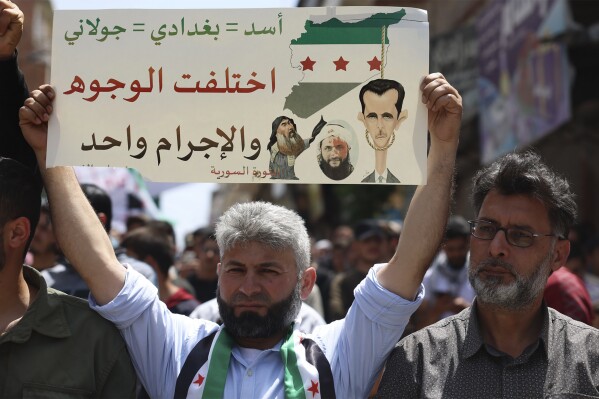 A Syrian protester holds up a placard during a protest against al-Qaida-linked Hayat Tahrir al-Sham group in Binnish town, in Idlib province, Syria, Friday, May 17, 2024. Members of Syria's most powerful insurgent group in the country's rebel-held northwest fired bullets in the air and beat up protesters with clubs Friday injuring some of them as weekslong protests demanding release of detainees and an end to the group's rule intensified. The placard shows drawings of Syrian President Bashar Assad, the late Islamic State group Abu Bakr al-Baghdadi and Abu Mohammed al-Golani leader of the Hayat Tahrir al-Sham, once linked to al-Qaida and the Arabic writing reads: "Assad = Baghdadi = al-Golani. The faces are different but the crime is one." (AP Photo/Ghaith Alsayed)