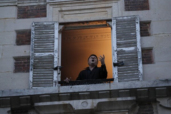 French tenor singer Stephane Senechal sings from his apartment window in Paris, Tuesday, March 24, 2020. French President Emmanuel Macron urged employees to keep working in supermarkets, production sites and other businesses that need to keep running amid stringent restrictions of movement due to the rapid spreading of the new coronavirus in the country. For most people, the new coronavirus causes only mild or moderate symptoms. For some it can cause more severe illness, especially in older adults and people with existing health problems. (AP Photo/Francois Mori)