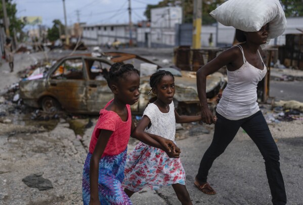 FILE - Girls holding hands are led past a burnt car blocking the street as they evacuate the Delmas 22 neighborhood to escape gang violence in Port-au-Prince, Haiti, May 2, 2024. As young Haitians are increasingly exposed to violence, the country is undergoing a wider push to dispel a long-standing taboo on seeking therapy and talking about mental health. (AP Photo/Ramon Espinosa, File)