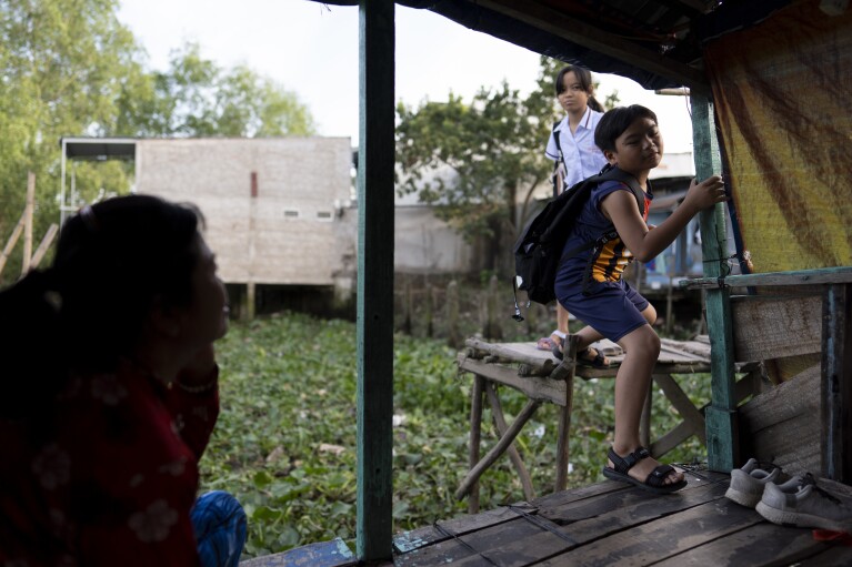 Nguyen Thi Thuy, left, sits on her houseboat as her twin grandchildren, Do Hoang Trung and his sister, Do Bao Tran, return from school in Can Tho, Vietnam, Wednesday, Jan. 17, 2024. After their mother left to pursue better financial opportunities in Ho Chi Minh City, the twins remained in the care of their grandmother, who supports the family by selling steamed buns at a floating market. (AP Photo/Jae C. Hong)